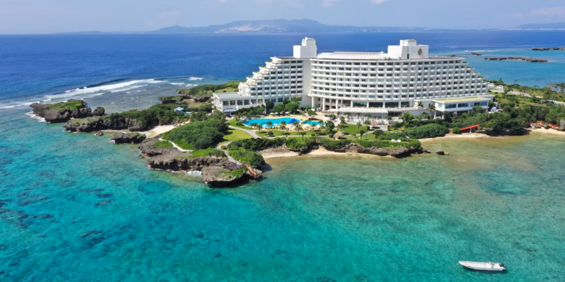 A large white hotel sits on a lush, rocky peninsula surrounded by clear turquoise ocean water, with a pool and gardens in front and a small boat in the foreground. An island is visible in the distance across the sea.
