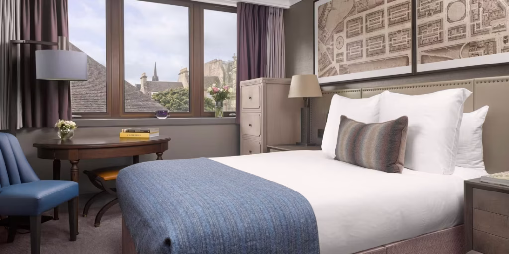 A cozy hotel room with a neatly made double bed, blue throw blanket, and accent pillow. There’s a round table with two chairs, large windows with city views, and framed maps above the bed.