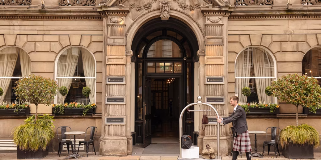 A hotel porter in a kilt stands outside an ornate stone building with arched windows, pushing a luggage cart. Small trees, plants, and outdoor tables with chairs line the entrance.