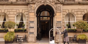 A hotel porter in a kilt stands outside an ornate stone building with arched windows, pushing a luggage cart. Small trees, plants, and outdoor tables with chairs line the entrance.