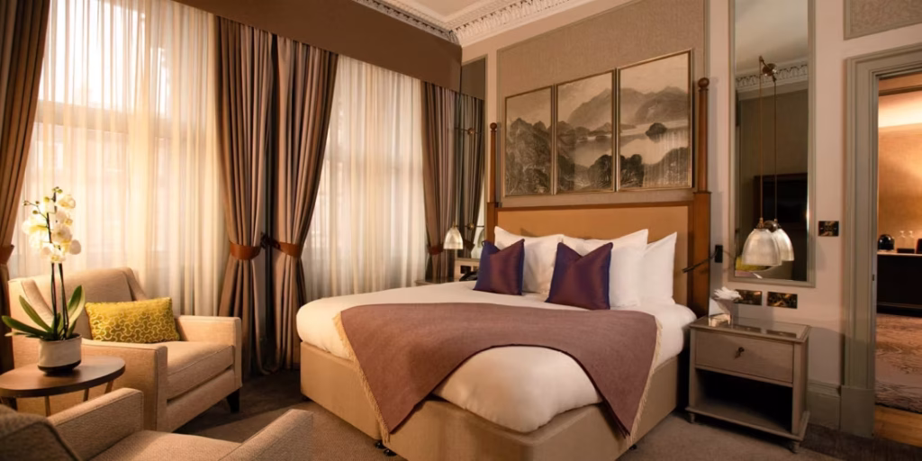 A stylish hotel room with a large bed, purple pillows, a mauve throw, elegant curtains, an armchair with a cushion, side tables, lamps, a potted orchid, and framed landscape art above the bed.