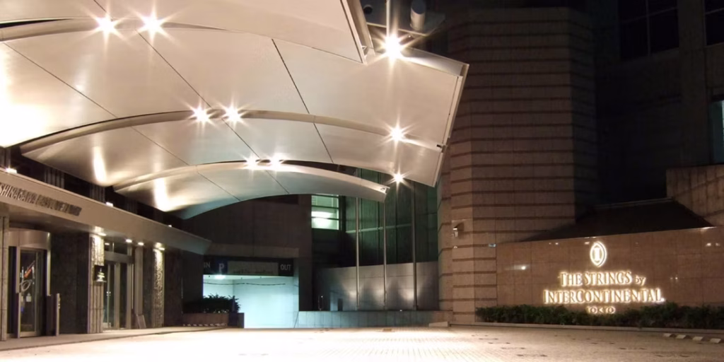 The entrance of The Strings by InterContinental Tokyo at night, featuring a modern, curved canopy roof and illuminated signage on a stone wall beside the glass doorway.