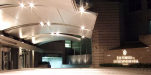 The entrance of The Strings by InterContinental Tokyo at night, featuring a modern, curved canopy roof and illuminated signage on a stone wall beside the glass doorway.