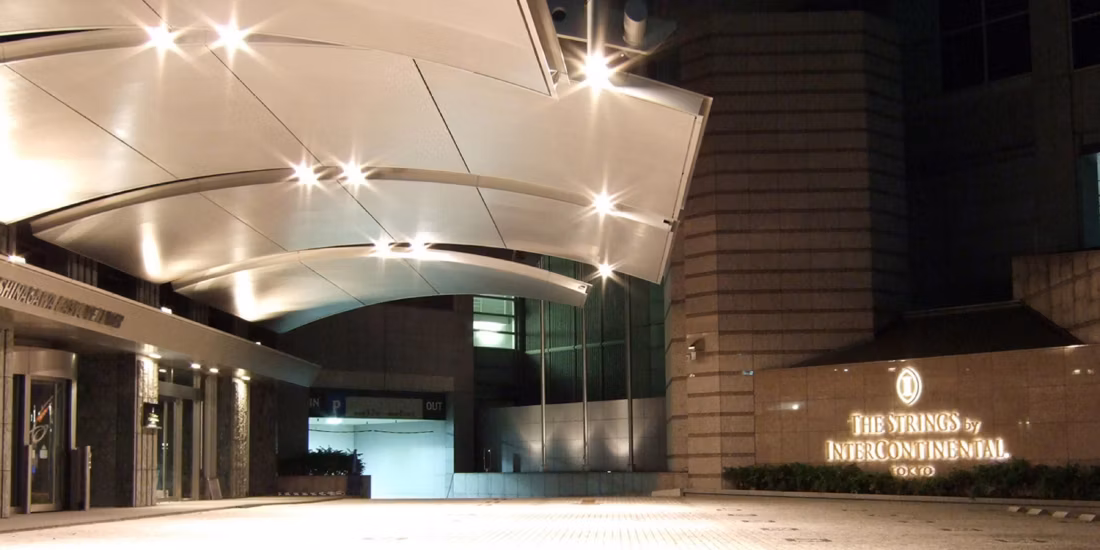 The entrance of The Strings by InterContinental Tokyo at night, featuring a modern, curved canopy roof and illuminated signage on a stone wall beside the glass doorway.