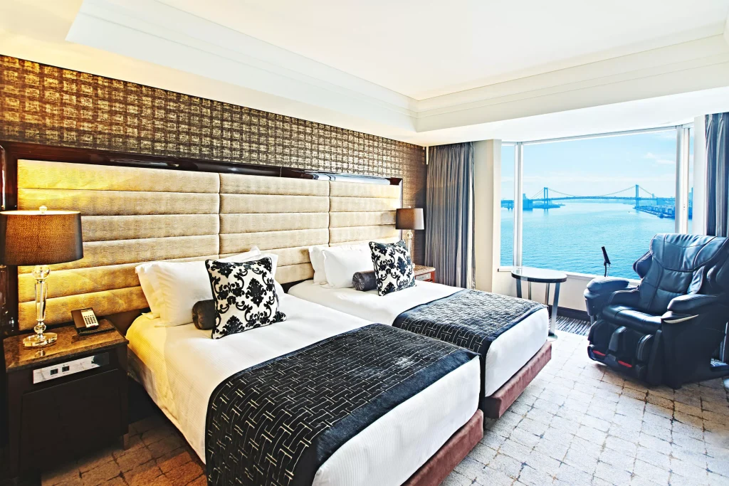 A hotel room with two twin beds, decorative pillows, bedside lamps, a massage chair, and large windows offering a view of a blue sea and a distant bridge.