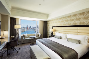 A spacious hotel room features a large bed with white and gray bedding, a sofa by a window, a desk with a chair, and lamps. The window offers a city skyline and river view under a bright blue sky.