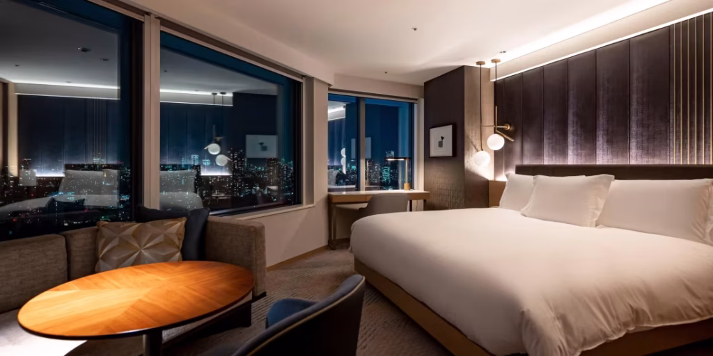 Modern hotel room at night with a large bed, corner sofa, round wooden table, desk, and floor-to-ceiling windows showing a city skyline illuminated with lights. Warm lighting creates a cozy and inviting atmosphere.