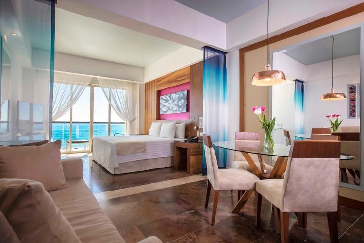 Modern hotel suite with a king bed, sofa, glass dining table with four chairs, and ocean view through large windows. The room features contemporary decor with fresh flowers and colorful artwork.