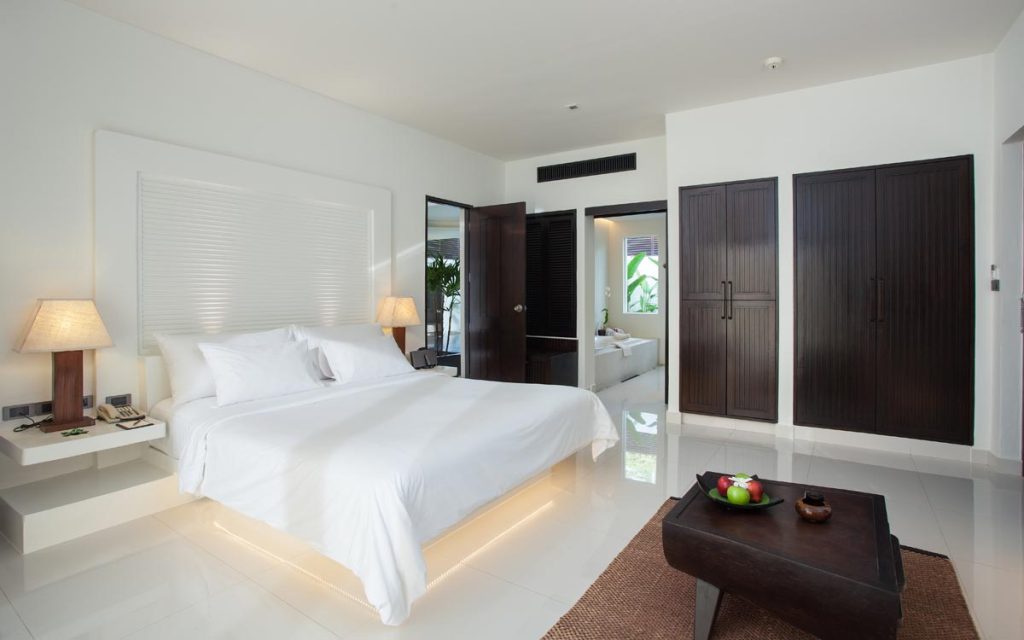 Modern hotel room with a large bed, white linens, bedside tables with lamps, dark wooden wardrobes, a small table with fruit, and an open door leading to a bathroom. The decor is minimalist and bright.