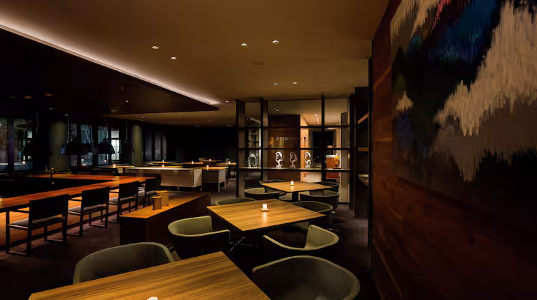 A modern, dimly lit restaurant with wooden tables, cushioned chairs, and soft overhead lighting. A colorful abstract painting hangs on the right wall, and candles on the tables create a cozy, inviting atmosphere.
