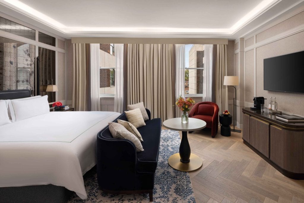 A modern hotel room with a large bed, dark blue sofa, round table with flowers, red armchair, flat-screen TV, and large windows with light curtains letting in natural light.