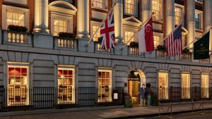 The exterior of The Beaumont hotel at night, warmly lit with large windows, columns, and multiple flags including the UK, Singapore, and US flags above the entrance, where a doorman stands.