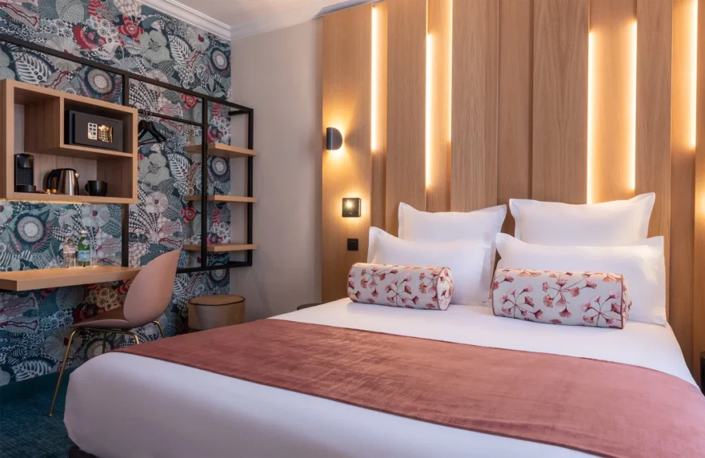 A stylish hotel room with a double bed, white linens, pink throw and bolster pillows, wooden headboard with vertical lights, a floral accent wall, a desk with a chair, and shelves holding a microwave, kettle, and safe.