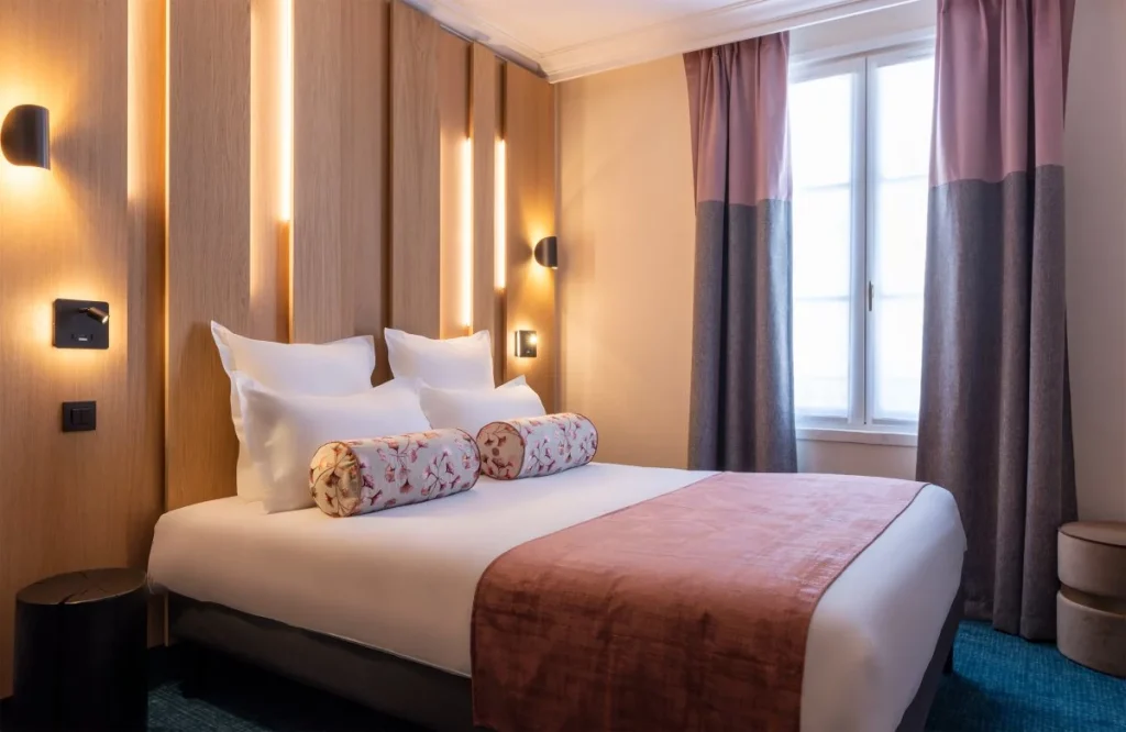 A modern hotel room with a double bed, white pillows, floral cylindrical cushions, and a pink throw blanket. The room features wood-paneled walls with built-in lights, curtains, and a large window letting in natural light.