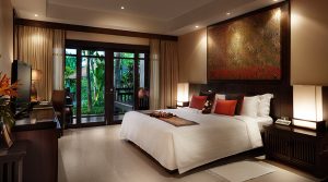 A modern hotel room with a large bed, red and brown pillows, bedside lamps, a desk, TV, and glass doors opening to a lush green garden. Warm lighting and a large artwork above the bed create a cozy atmosphere.