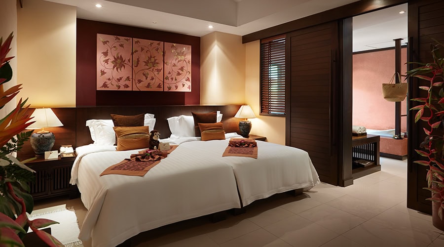 A stylish hotel room with two twin beds topped with white linens and brown accent pillows, side tables with lamps, floral artwork above the beds, and an adjoining space with a modern bathtub.