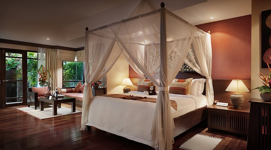 A spacious, elegant bedroom with a four-poster canopy bed, warm lighting, wooden floors, bedside tables with lamps, a seating area, large windows, and tropical decor.