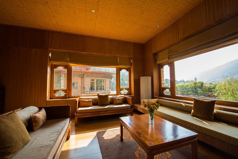 A cozy living room with wooden walls and ceiling, large windows letting in sunlight, cushioned sofas, a coffee table with flowers, and scenic mountain views outside.