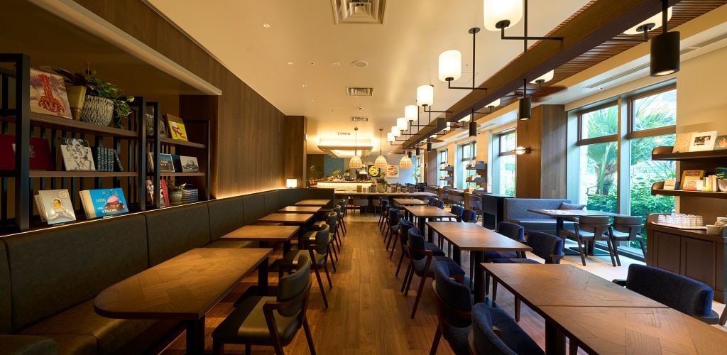 A modern, well-lit café with wooden tables and chairs, bookshelves along one wall, large windows letting in natural light, and decorative light fixtures hanging from the ceiling.