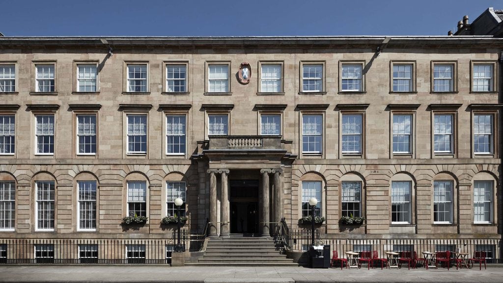 A large, symmetrical stone building with three floors, many windows, a central columned entrance, and steps leading up to the door. Red chairs and tables are arranged outside on the right side.