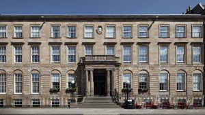 A large, symmetrical stone building with three floors, many windows, a central columned entrance, and steps leading up to the door. Red chairs and tables are arranged outside on the right side.