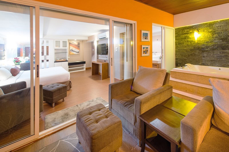 Hotel suite with a cozy sitting area, ottomans, and table in the foreground, a hot tub on the right, and a bedroom with a large bed, TV, and modern decor visible through sliding glass doors.