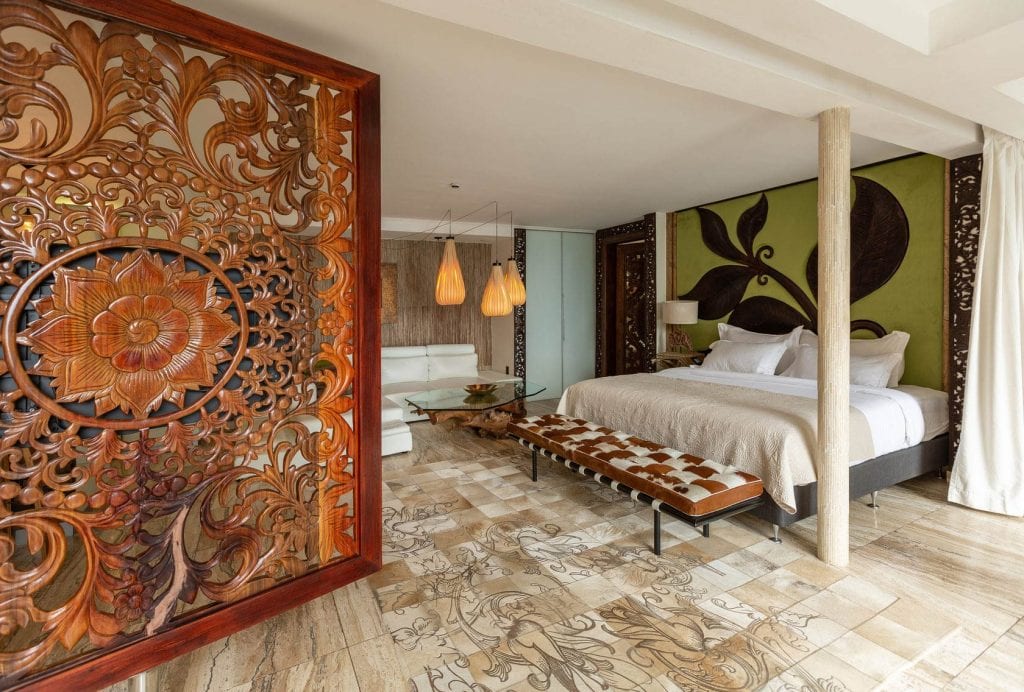A spacious, stylish bedroom with a king-size bed, carved wooden room divider, patterned tile floor, green accent wall with floral art, modern furniture, and pendant lights. Natural light fills the space.