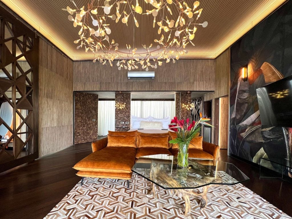 A luxurious modern living room features a brown velvet sectional sofa, glass coffee table, geometric rug, and artistic light fixtures. The space has wood-paneled walls, large mirrors, and a floral arrangement on the table.