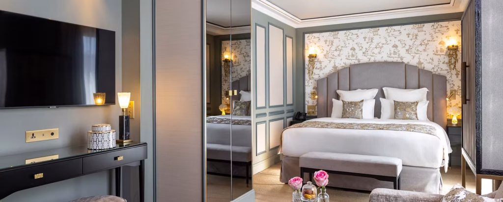 Elegant hotel room with a large bed, plush pillows, and a gray upholstered headboard. There are wall sconces, a flat-screen TV, mirrored closet doors, a black desk, and fresh pink flowers on a bench at the foot of the bed.