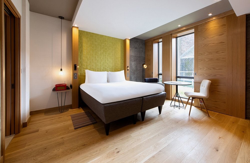 A modern hotel room with wooden floors, a large bed with white bedding, a green accent wall, a small round table with two chairs by the window, and warm lighting. A red phone sits on a side table.