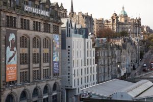 View of historic stone buildings in Edinburgh, Scotland, including the City Art Centre with art banners and the modern Market Street Hotel, with a mostly empty street and distant classical architecture.