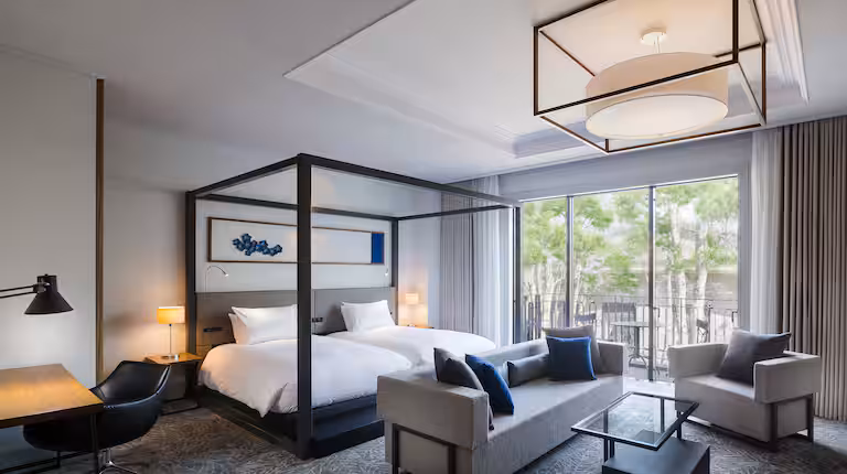Modern hotel room with a large four-poster bed, a gray sofa, desk with chair, and floor-to-ceiling windows leading to a balcony overlooking greenery. The room is decorated in neutral tones with blue accents.
