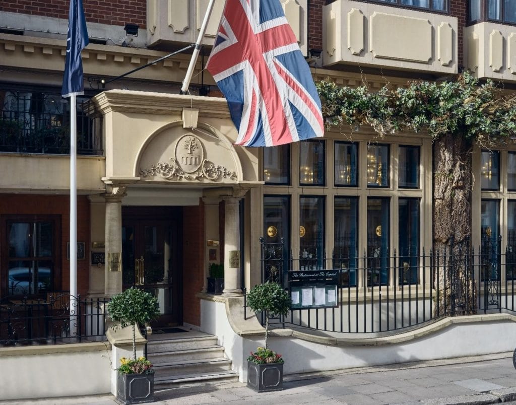 Entrance to a building with large windows, decorative columns, potted plants, and a Union Jack flag overhead. There is a wrought-iron fence and greenery above the windows.