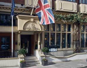 Entrance to a building with large windows, decorative columns, potted plants, and a Union Jack flag overhead. There is a wrought-iron fence and greenery above the windows.