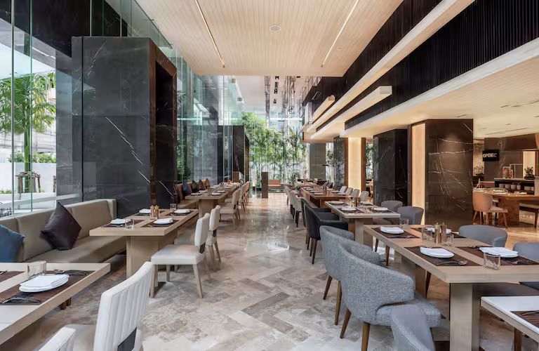 Modern, spacious restaurant interior with neatly arranged wooden tables and cushioned chairs, large floor-to-ceiling windows, marble floors, and abundant natural light. Greenery is visible outside the windows.