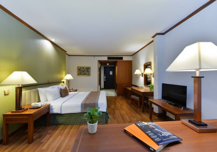 A spacious hotel room with a large bed, wooden furniture, bedside lamps, a TV, a desk with a plant and magazines, and warm lighting. The floor is wooden, and there is artwork on the far wall near the door.