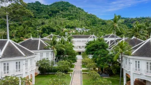 White colonial-style buildings with grey roofs surround a central walkway, lush greenery and palm trees fill the courtyard, and a forested hill rises in the background under a blue sky.