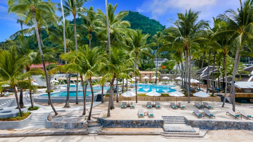 A tropical resort features a large pool surrounded by palm trees, sun loungers, and white umbrellas, with a lush green mountain and blue sky in the background.