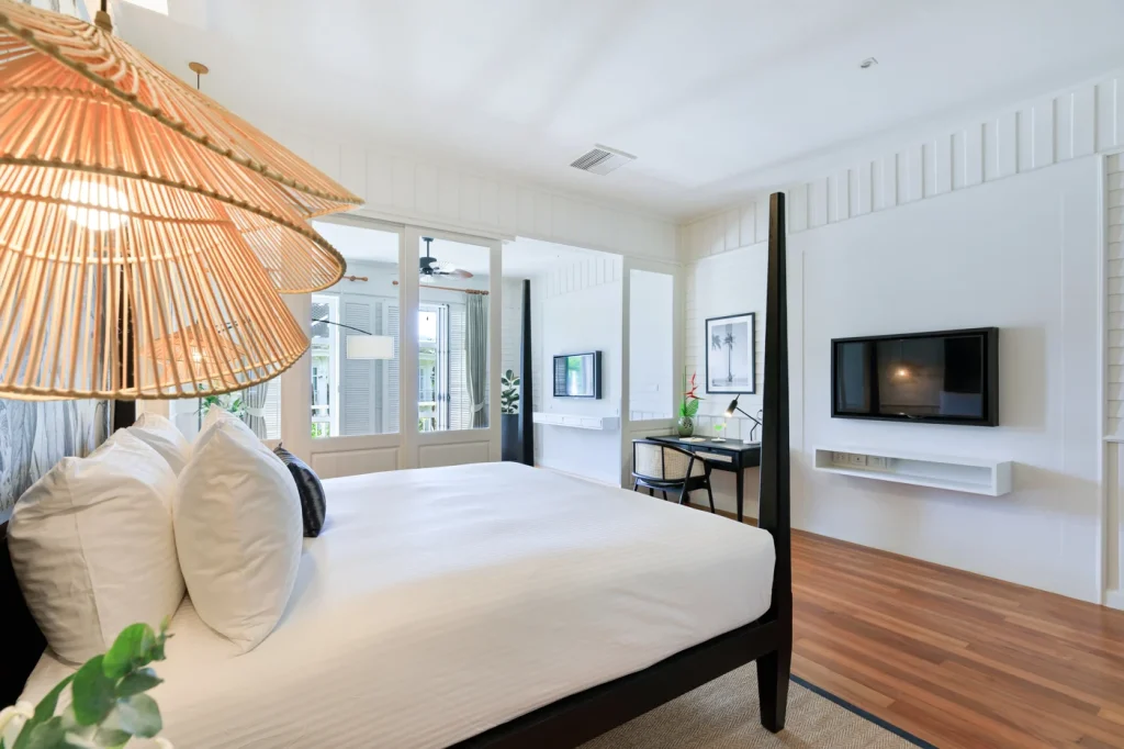 Bright, modern bedroom with a large bed, white bedding, wood floors, a wall-mounted TV, desk, and woven pendant lights. Large windows let in natural light and overlook a balcony with greenery visible outside.