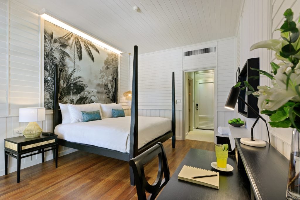 A modern hotel room with a four-poster bed, tropical mural, wooden floors, a desk with a notepad and drink, and a vase of white lilies. The room is bright with white walls and a bathroom entrance visible.