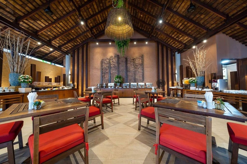 A spacious, elegant restaurant with wooden tables and chairs featuring red cushions, decorated with plants and flowers, and a buffet setup along the back wall under a high, wooden ceiling.