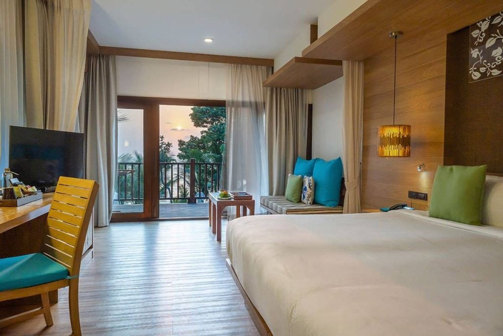 A modern hotel room with a large bed, desk, TV, and a seating area. Sliding glass doors open to a balcony with a view of trees and a sunset sky. Soft lighting and wood accents create a cozy atmosphere.