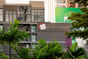 A modern gray building with large windows displays a Hotel Clover Asoke sign. Green leafy plants are in the foreground, and colorful tall buildings are visible in the background.
