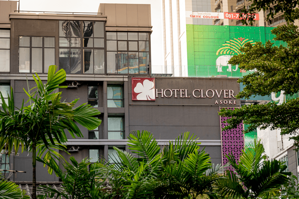 A modern gray building with large windows displays a Hotel Clover Asoke sign. Green leafy plants are in the foreground, and colorful tall buildings are visible in the background.