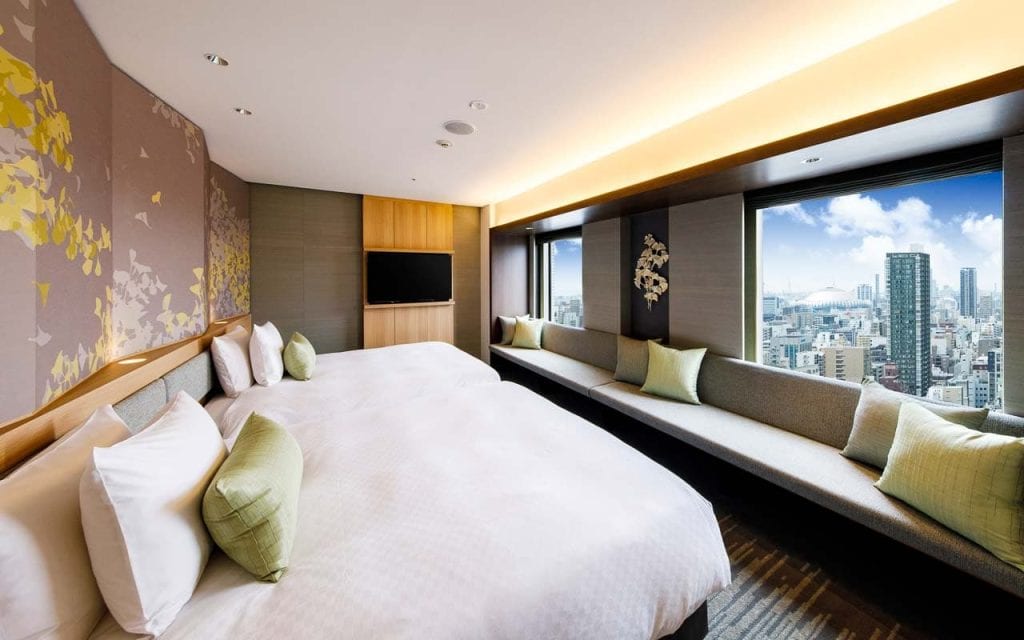 Modern hotel room with two large beds, a long cushioned bench by wide windows, decorative pillows, a wall-mounted TV, and city skyline views under a bright, partly cloudy sky.