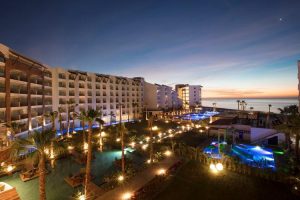 A modern beachfront resort at dusk, with illuminated pools, palm trees, and multi-story buildings overlooking the ocean under a colorful evening sky.