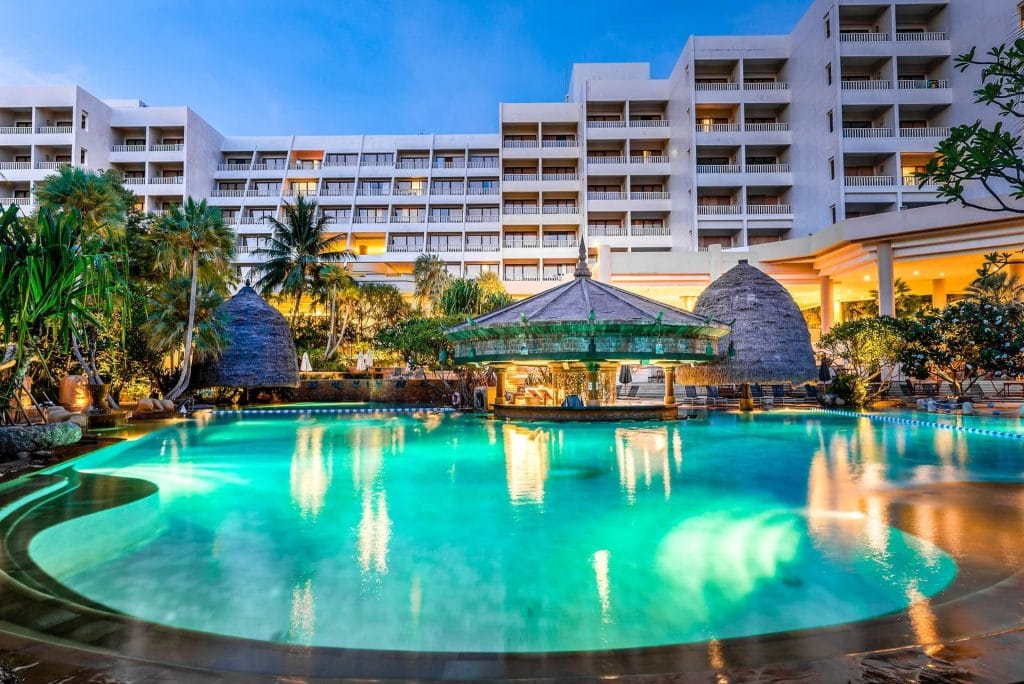 A large, illuminated outdoor swimming pool with a swim-up bar is surrounded by tropical plants in front of a multi-story modern hotel building at dusk.