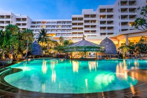 A large, illuminated outdoor swimming pool with a swim-up bar is surrounded by tropical plants in front of a multi-story modern hotel building at dusk.