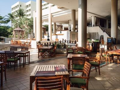 Outdoor patio of a modern hotel or resort with wooden tables and chairs, surrounded by palm trees and columns, with natural sunlight and a relaxed atmosphere.