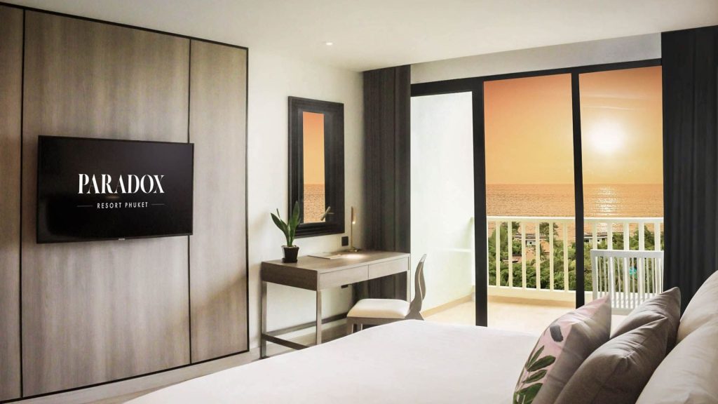 A modern hotel room with a bed, desk, wall-mounted TV displaying PARADOX Resort Phuket, and a balcony overlooking the ocean at sunset.